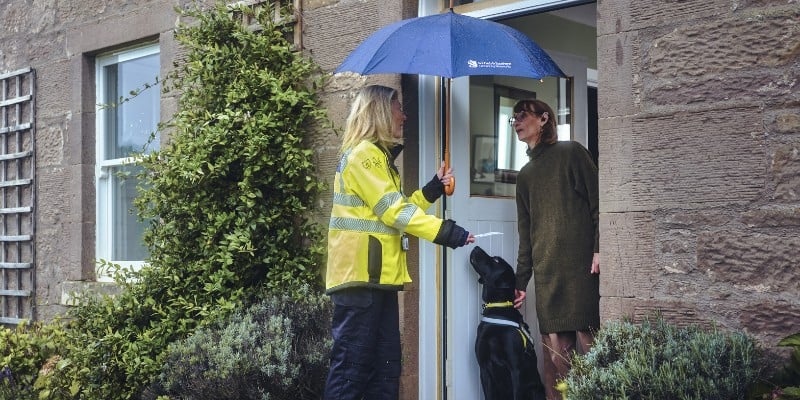Woman in high-vis with umbrella hands item to person at doorway with a dog.