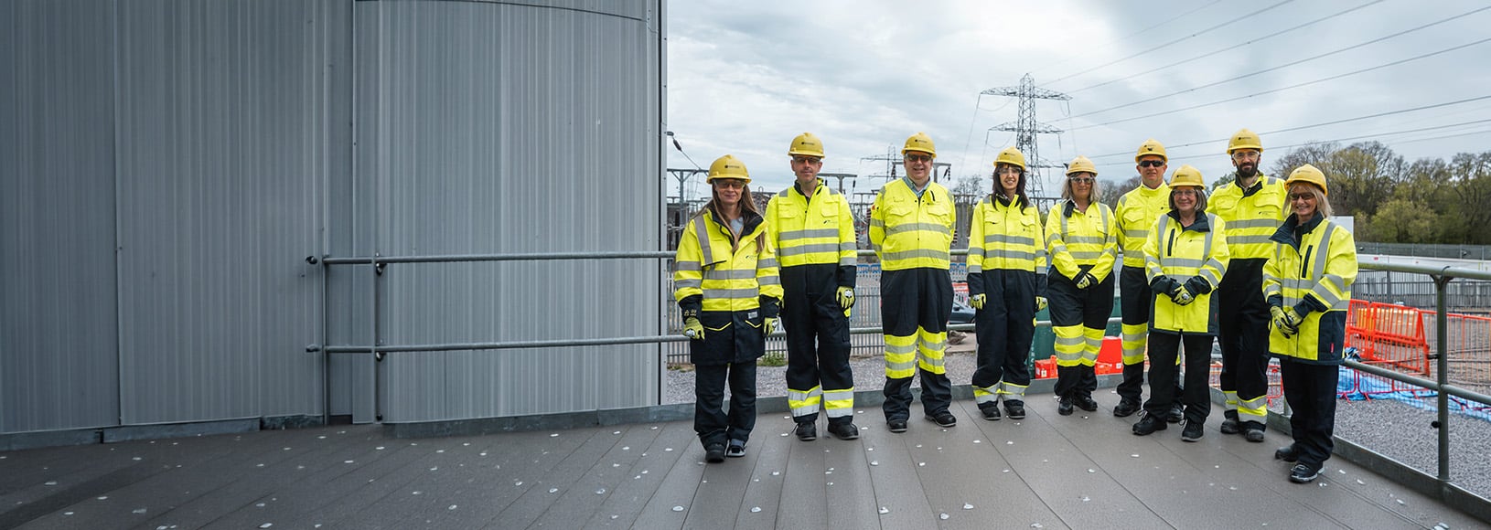Nine people in yellow hard hats and high-vis jackets stand outdoors.