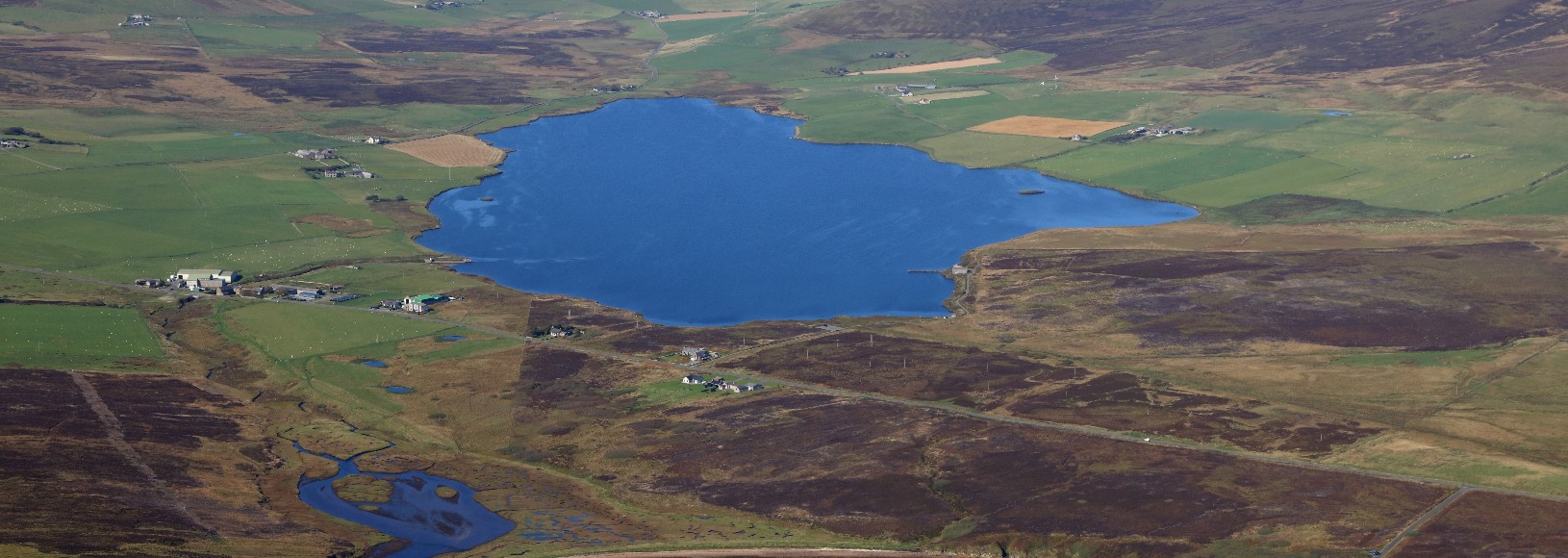 Aerial view shows a blue lake bordered by green fields, brown moorland, and clustered buildings.