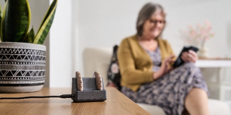 Pair of hearing aids charging on a table; a blurred woman uses a device.