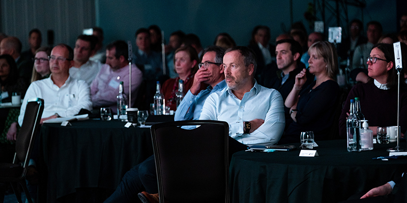Image of people in presentation room listening to speaker on stage 