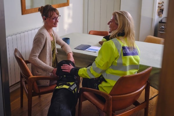 Two women sit at a table petting a black dog wearing a working harness.