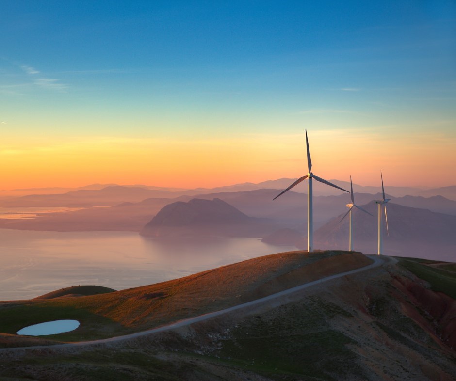 Scenic view of wind turbines on a mountain ridge at sunset, overlooking a calm sea and distant islands, symbolising clean energy and environmental sustainability.
