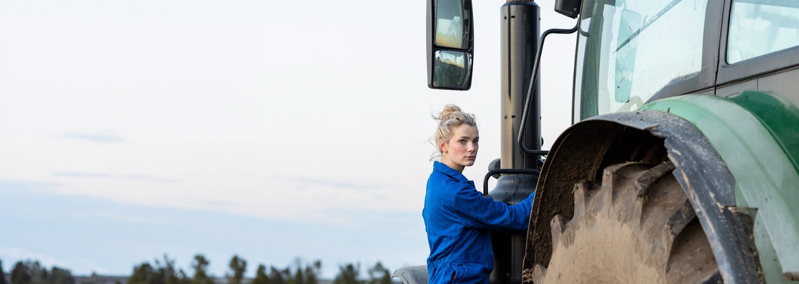 Woman in blue overalls getting into a green tractor.